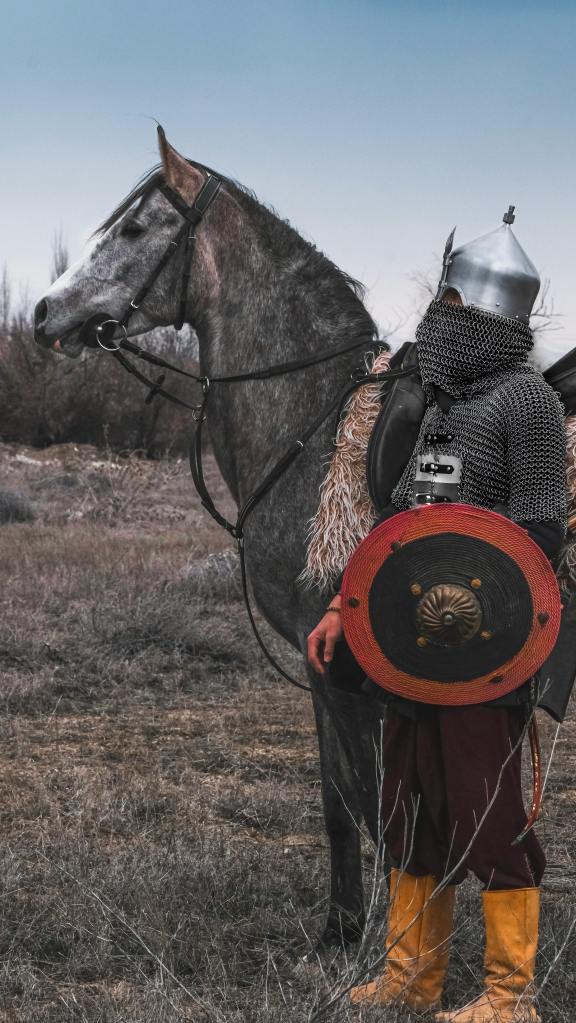 Turkish soldier in armor holding a shield standing next a horse.